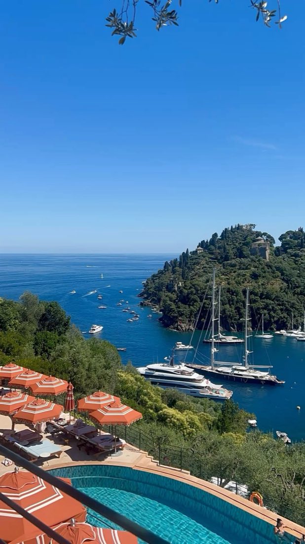 View over a rocky bay on the Italian Riviera, yachts moored below a pool lined with orange umbrellas
