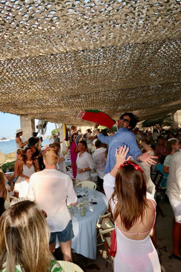 A crowded beach club lunch under a shade canopy on the French Riviera, guests dressed in white for the occasion